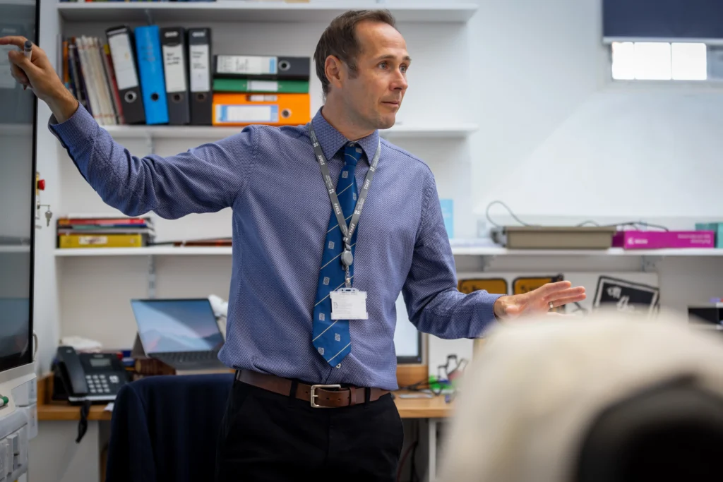A teacher at The King's Summer School International Relations Academy guiding his students through a lesson. he is wearing clear identification and using a large flatscreen monitor for visual effect in teaching.